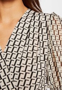 Zavinovací blůza vyrobená z lehkého materiálu, s krémovým pozadím a geometrickým černým vzorem. Rukávy jsou průhledné s plošnými detaily.
