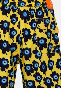 Pantaloni corti gialli con grandi fiori blu navy e dettagli blu al centro. Presentano una tasca laterale e una cintura elastica. Tessuto dalla texture liscia.