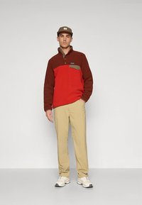 Rød og brun fleecepullover med lomme med grønne detaljer, beige bukser og brun kasket. Hvide sneakers fuldender outfittet.