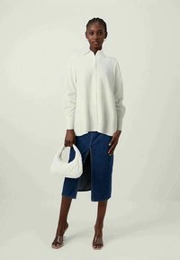 Camisa branca de botão com mangas longas, combinada com uma saia de ganga de comprimento médio com uma fenda frontal; modelo segurando uma mala branca acolchoada.