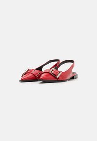 Červené kožené slingback topánky s špičatou špičkou, zdobené striebornými prackami a kruhovými výrezmi, na plochej čiernej podrážke.