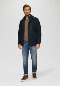 Marineblauer Wollmischmantel mit einer mit Reißverschluss versehenen Brusttasche, kombiniert mit einem braunen Pullover, hellblauen Jeans und braunen Sneakern. Glatte Textur.