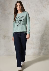 Hellgrünes Langarmshirt mit grafischem Textdesign, kombiniert mit dunkelblauen Straight-Leg-Jeans und weißen Sneakers. Weiches Material und lässige Passform.