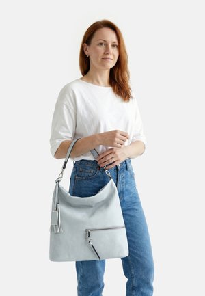 Femme aux cheveux roux portant un haut blanc et un jean bleu, tenant un grand sac à main gris clair avec un pompon et une poche zippée à l'avant.