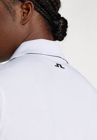 Camisa polo branca com gola texturizada, apresentando um detalhe de logotipo preto nas costas e um pequeno piping preto ao longo da borda da gola.