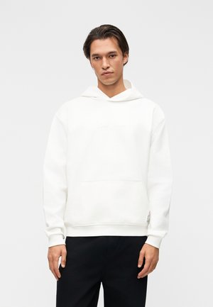 Young man wearing a plain white hoodie with a front pocket and black pants, standing against a light gray background.