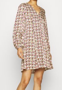 Femme portant une robe florale à manches longues, amples, avec des fleurs roses, jaunes et noires sur un fond blanc, se tenant devant un arrière-plan uni.