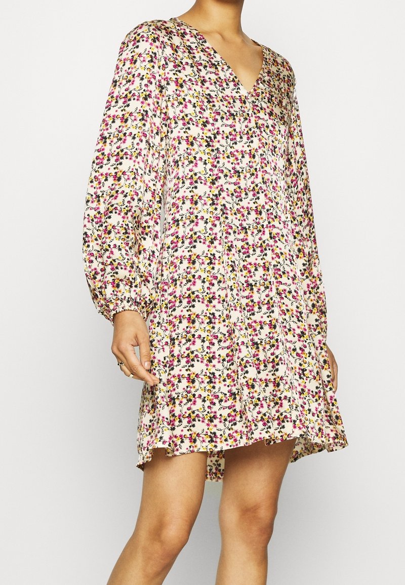 Femme portant une robe florale à manches longues, amples, avec des fleurs roses, jaunes et noires sur un fond blanc, se tenant devant un arrière-plan uni.