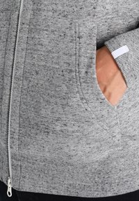 Sudadera gris con cremallera y tejido texturizado y moteado. Presenta bolsillos laterales y puños y dobladillo de canalé, con un pequeño acento de etiqueta blanca.