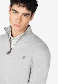 Suéter gris de punto con cuello falso y media cremallera. Presenta un bordado del logo en azul marino en el pecho. Textura suave, diseño ajustado.