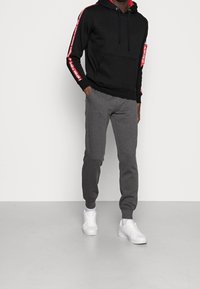 Sudadera negra con capucha y acentos rojos, combinada con pantalones jogger grises y zapatillas blancas. El modelo está de pie en un fondo neutro.