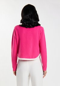 Maglione corto in pile di un vivace colore rosa, con una texture morbida, caratterizzato da un orlo arrotondato e un bordo contrastante bianco. Maniche lunghe con polsini aderenti.