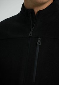 Černá fleecová bunda s vysokým límcem, která má dvě zipové kapsy a trojúhelníkové táhla zipu. Hladká textura, minimalistický design.