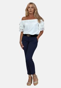 Biała bluzka typu off-the-shoulder z warstwowymi falbanami, połączona z granatowymi spodniami w paski i beżowymi sandałami na platformie. Czarny pasek z złotą klamrą.