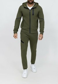 Survêtement sportif vert olive avec veste  à capuche zippée et pantalon ajusté comportant des accents noirs, fabriqué en tissu lisse et extensible. Baskets blanches.