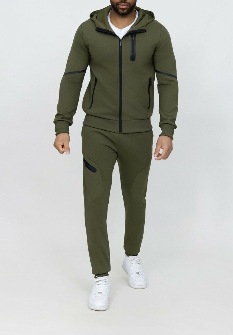 Survêtement sportif vert olive avec veste à capuche zippée et pantalon ajusté comportant des accents noirs, fabriqué en tissu lisse et extensible. Baskets blanches.