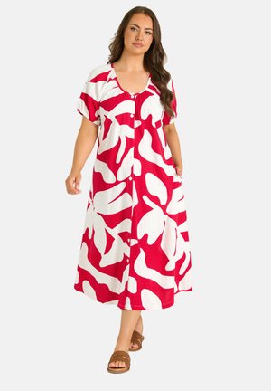 Kvinde iført en rød og hvid midi-kjole med abstrakt mønster, korte ærmer og brune sandaler, stående foran en ensfarvet baggrund.