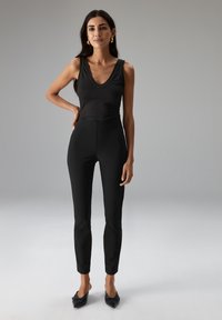 Zwarte geribbelde mouwloze top gecombineerd met nauwsluitende zwarte broek, beide gemaakt van gladde stof. Opvallende zwarte mules met puntige neus maken de look compleet.