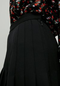 Zwarte plooienrok met een gladde textuur, voorzien van een aansluitende tailleband en subtiele stikdetails, gecombineerd met een blouse met bloemenpatroon.