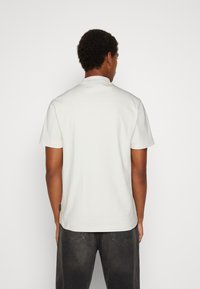 White short-sleeve t-shirt with a round neck, cotton fabric, and a tag on the left side near the hem, paired with black jeans.
