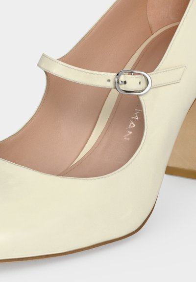 Chaussure Mary Jane couleur crème avec une bride à boucle argentée et un petit talon carré sur fond blanc.