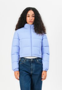 Lys lilla kort pufferjakke med høy krage, horisontalt quiltet design og frontglidelås; kombinert med mørkeblå jeans.