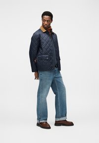 Μπλε καπιτονέ μπουφάν με καφέ γιακά, δύο μπροστινές τσέπες και κλείσιμο με τρουκ, συνδυασμένο με ανοιχτό μπλε τζιν παντελόνι με ίσια γραμμή και καφέ παπούτσια.