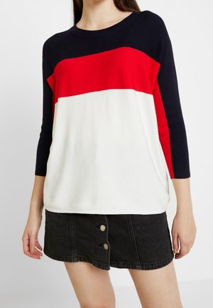 Femme portant un pull colorblock noir, rouge et blanc avec des manches trois-quarts et une mini-jupe en denim noir avec des boutons sur le devant.