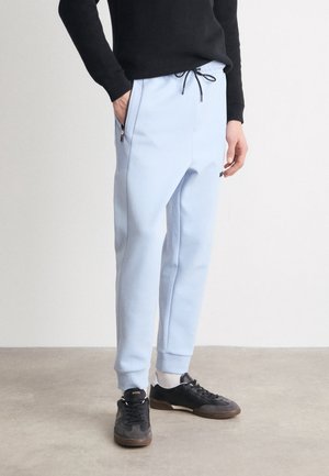 Personne portant un pantalon de jogging bleu clair avec des poches zippées, un pull noir, des chaussettes blanches et des baskets noires, debout sur un sol en béton.