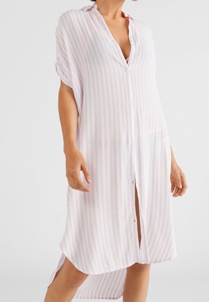 Mujer que lleva un vestido camisa suelto, hasta la rodilla, de manga corta y con botones, con rayas verticales de color rosa claro.
