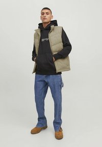 Olívovo zelená vesta s výplňou cez čiernu mikinu s kapucňou, doplnená svetlomodrými džínsami a hnedými čižmami. Neformálne oblečenie s funkčným vrstvením.