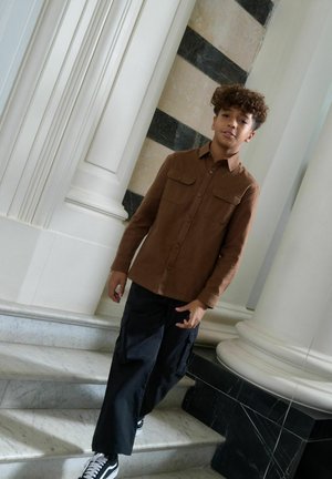 Chemise marron à boutons avec poches poitrine, associée à un pantalon cargo noir et des baskets noires. Le modèle se tient sur des marches en marbre devant un arrière-plan de colonnes.
