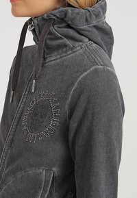 Hoodie zippé gris en tissu doux, avec un col montant et un logo de crâne en rhinestones décoratif sur la poitrine. Deux poches latérales incluses.