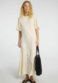 Vestido de color crema, de manga larga, con detalles de costuras diagonales. Tejido suave, ajuste holgado. La modelo sostiene un bolso de punto negro. Zapatos planos negros.