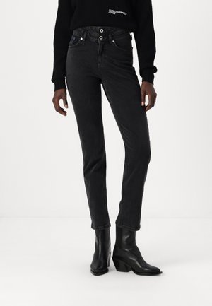 Schwarze hochtaillierte Jeans mit schlankem Schnitt, ausgestattet mit zwei vorderen Knöpfen, fünf Taschen und einer dezenten, ausgeblichenen Textur. Kombiniert mit schwarzen Absatzstiefeln.