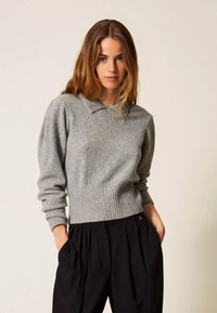 Maglione grigio con orlo a coste e maniche lunghe, caratterizzato da uno scollo a collo con perline. Abbinato a pantaloni neri plissettati.
