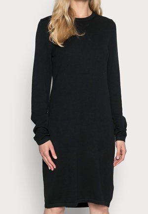 Kobieta o długich blond włosach, ubrana w prostą czarną sukienkę z długim rękawem sięgającą kolan, stojąca na neutralnym tle.