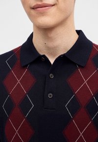 Gros plan d'une personne portant un pull à motif losanges bleu marine et bordeaux avec un col et deux boutons.
