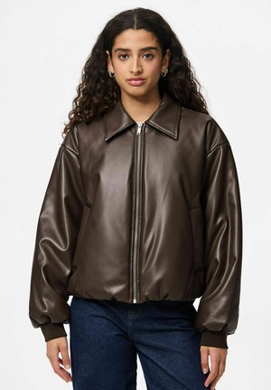 Femme aux longs cheveux bouclés portant une veste en cuir marron zippée et un jean bleu foncé, faisant face vers l’avant avec une expression neutre.
