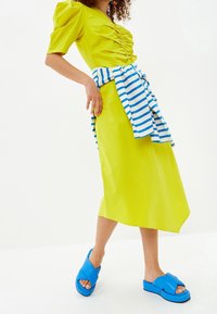 Limetkově zelené šaty s nařasením, se zeleno-bílým pruhovaným topem a doplněné velkými modrými sandály. Hladký povrch, střední délka.