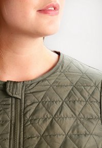Olive green quilted jacket with diamond pattern, featuring a zip closure and rounded neckline. Made of soft, smooth fabric.