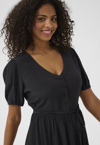 Robe noire avec manches bouffantes, décolleté en V, boutons avant et taille nouée. Fabriquée en tissu texturé, elle présente une jupe fluide.