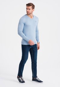 Hellblaues, langärmliges V-Ausschnitt-Shirt mit gerippten Bündchen, kombiniert mit dunkelblauen Slim-Fit-Jeans und marineblauen Sneakers mit weißen Schnürsenkeln.
