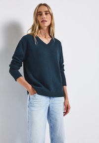 Donkergroene gebreide trui met een V-hals, lange mouwen en ribbels aan de manchetten, gecombineerd met lichtblauwe spijkerbroek.