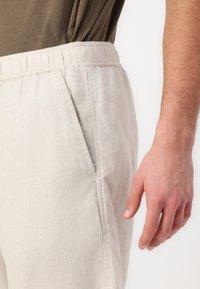 Lys beige linbukser med elastisk linning, sidelommer og en glat tekstur. Stoffet er let og åndbart.