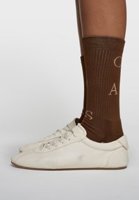 Weiß strukturierte Lederschuhe mit Schnürsenkeln. Braune, gerippte Socken mit hellem Text, über dem Knöchel getragen. Einfaches, minimalistisches Design.