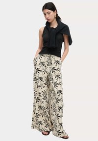 Zwarte mouwloze top met een strik bij de hals, gecombineerd met crèmekleurige broek met wijde pijpen en een zwart bloemenpatroon. Er worden zwarte sandalen gedragen.