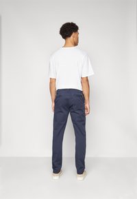 T-shirt bianca a maniche corte abbinata a pantaloni blu scuro, con tasche laterali e una texture liscia, vista da dietro.