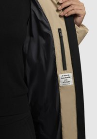 Sort yderstofjakke med en beige foret inderside. Har en lynlås lomme og et mærke med teksten "Et varmt kram lavet af stof."