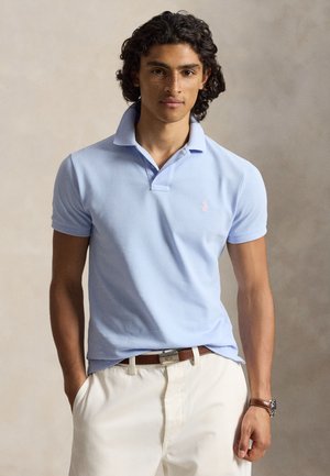 Młody mężczyzna z kręconymi włosami, ubrany w jasnoniebieską koszulkę polo, beżowe spodnie, brązowy pasek i zegarek na nadgarstku, stojący z jedną ręką w kieszeni.